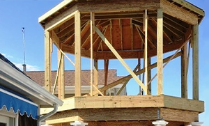 A wooden gazebo under construction, featuring an octagonal shape with exposed beams and a partially completed roof. The sky is clear and blue.