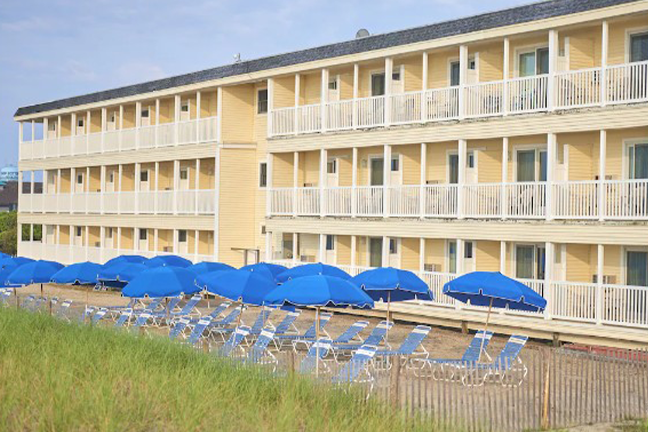 Yellow three-story hotel with white balconies. In front, a row of blue beach umbrellas and lounge chairs on a sandy area. Bright, sunny atmosphere.