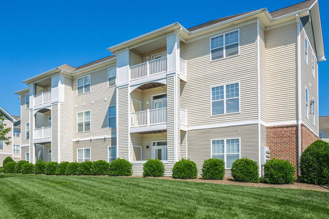 Three-story beige apartment building with white trim and balconies, set against a clear blue sky. Manicured green lawn and shrubs in the foreground. Calm, sunny day.