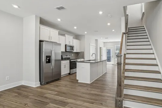 A modern kitchen with stainless steel appliances, white cabinets, and a center island. Hardwood floors and a staircase create a warm, inviting feel.