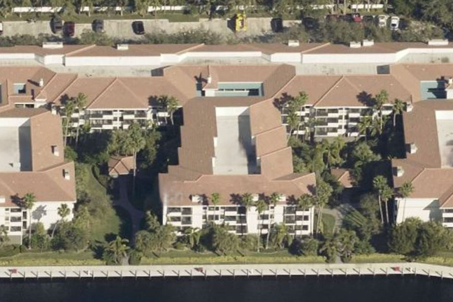 Aerial view of a beige building with a red-tiled roof, surrounded by greenery and located near a calm waterway, creating a serene atmosphere.
