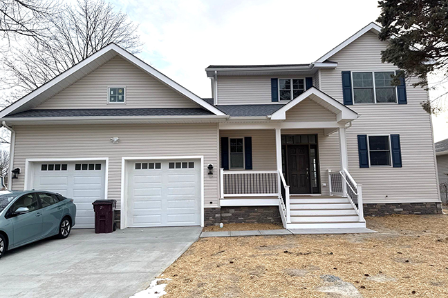 A two-story house with a beige exterior and dark shutters, featuring a double garage and a front porch with steps. The lawn is dry, and the sky is overcast.