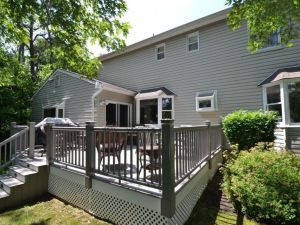 A cozy gray wooden deck with railing attached to a house. Set with outdoor furniture, a grill, surrounded by lush greenery and trees, conveying tranquility.