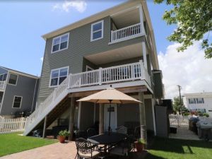 Three-story house with gray siding and white trim, featuring upper balconies and stairs leading to a patio. A table with an umbrella sits below, conveying a relaxed outdoor setting under a clear blue sky.