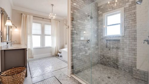 Luxurious bathroom with a glass-enclosed shower, gray tile walls, marble floors, and a sunlit soaking tub area. Elegant and serene ambiance.